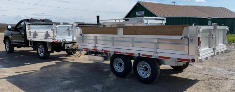 A heavy-duty pickup truck hitched to a large aluminum dump trailer with dual axles and wooden side extensions, parked on a paved surface near a green-roofed building.
